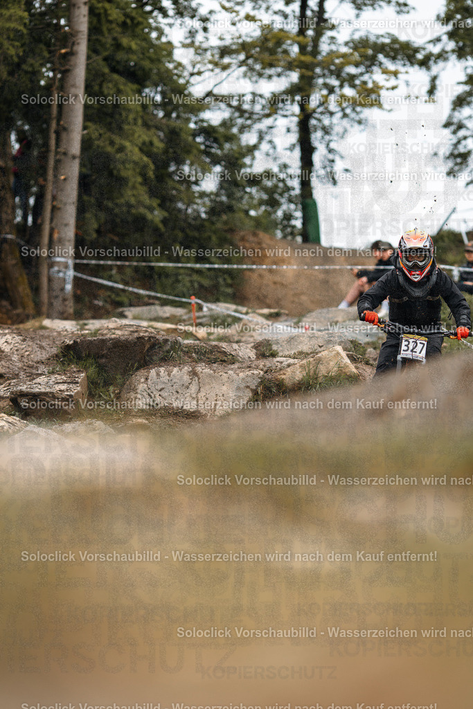 Mountainbike-Action im Gelände | Das Foto zeigt einen Mountainbiker in voller Montur, der mit hoher Geschwindigkeit eine staubige Kurve auf einem anspruchsvollen Trail durchquert. Der Fahrer trägt einen schwarzen Helm mit roten Akzenten und eine dunkle Brille. Er trägt eine schwarze Jacke und Hose. Der Staub, der von den Reifen aufgewirbelt wird, verleiht dem Bild eine dynamische Atmosphäre. Im Hintergrund sind Bäume und eine rote Markierung sichtbar. - Realisiert mit Pictrs.com