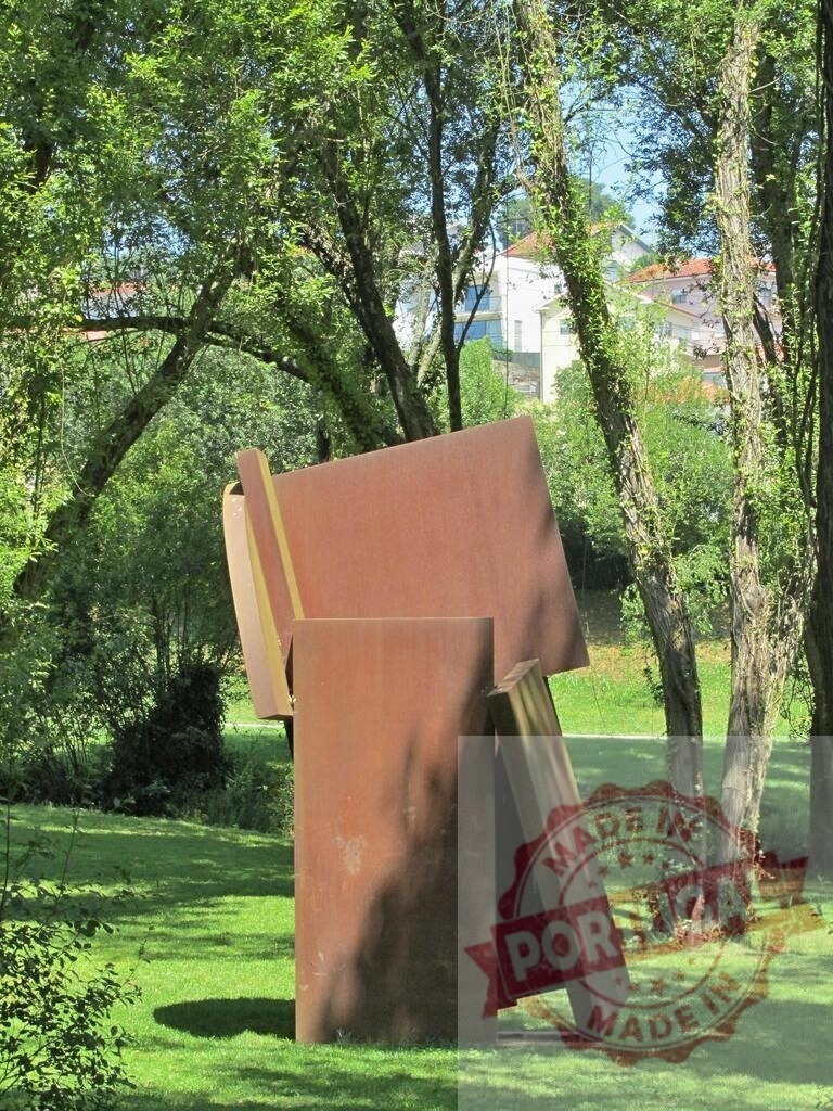 Abstract rusted metal sculpture in green park landscaping. Portugal - Norte | Abstract corten steel sculpture standing in a sunny public park with green grass and trees - Realized with Pictrs.com