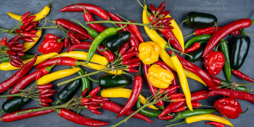 Farbenexplosion: Bunte Chilischoten auf Schieferplatte | Ein lebendiges Farbenspiel verschiedener Chilischoten erstreckt sich über den schwarzen Schiefer Hintergrund. Leuchtend rote, gelbe und tiefschwarze Schoten vermischen sich mit grünen Exemplaren zu einer beeindruckenden Gewürzkomposition. Kleine rote Chilisträuße kontrastieren mit länglichen gelben und schwarzen Sorten. Die glänzenden Oberflächen der frischen Schoten reflektieren das Licht und verstärken die Farbintensität. Diese kunstvolle Anordnung zeigt die natürliche Vielfalt scharfer Gewürze und inspiriert zu kreativen Kochexperimenten mit verschiedenen Schärfegraden. - Realisiert mit Pictrs.com