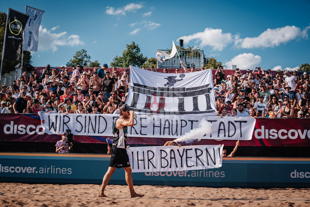 Beachvolleyball | Männer | Allianz German Beach Tour 2025 | Tourstop München | 13.07.2025 | Paul Henning läuft ein in das Stadion während die Fangruppe Fraktion Bräune eine Choreo mit Pyrotechnik zeigt. Wir sind eure Hauptstadt ihr Bayern