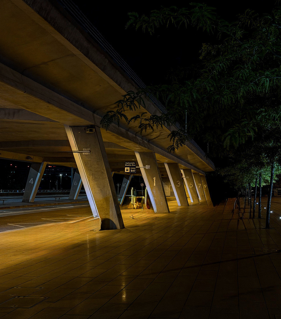 Midnight Brutalism – Erleuchtete Betonarchitektur am EuroAirport | Bringen Sie die rohe, kraftvolle Ästhetik moderner Betonarchitektur in Ihre Räume. Diese kunstvolle Nachtaufnahme fängt das Abflugterminal des EuroAirports um Mitternacht in einer beeindruckenden Licht- und Schattensymphonie ein. Die markanten, diagonalen Betonpfeiler und die strukturierte Deckenkonstruktion werden durch das warme Kunstlicht perfekt inszeniert, während die Dunkelheit der Nacht dem Motiv eine geheimnisvolle, urbane Tiefe verleiht. Ein exklusives Kunstwerk im Industrial- und Brutalismus-Stil, das in modern reduzierten Wohnzimmern, stylischen Lofts oder repräsentativen Geschäftsräumen ein absolutes visuelles Highlight setzt.• Material-Empfehlung: Für diese kontrastreiche Nachtaufnahme mit Betonstrukturen empfehle ich dieses Motiv dringend auf mattem Alu-Dibond. Es verhindert störende Lichtreflexionen in den tiefen Schwarztönen und unterstreicht den rauen, industriellen Charakter des Betons perfekt.• Call-to-Action: Setzen Sie ein kompromissloses Design-Statement an Ihrer Wand – wählen Sie jetzt Ihre Wunschgröße und bestellen Sie dieses exklusive Kunstwerk.[Archiv-Nr. #0108] - Realisiert mit Pictrs.com