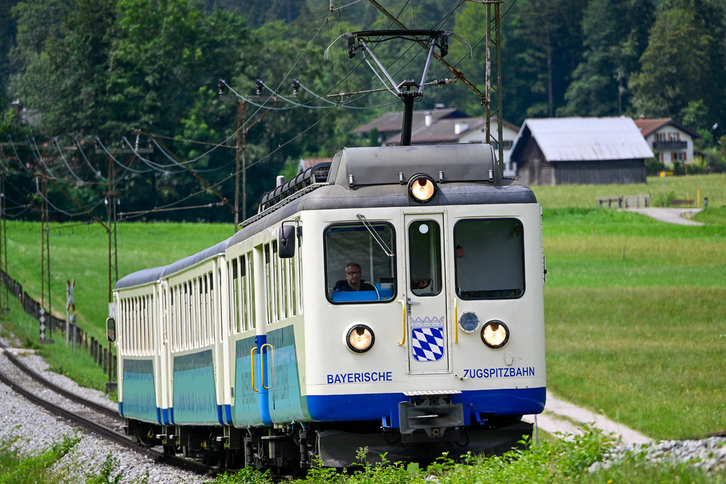 Sehnsucht auf Schienen: Die Reise zum Gipfel | Ein Anblick, der pure Nostalgie, Vorfreude und ein tiefes Heimatgefühl weckt: Mit eingeschalteten Scheinwerfern bahnt sich der klassische Triebwagen der Bayerischen Zugspitzbahn seinen Weg durch die idyllische Alpenlandschaft. Das charakteristische weiß-blaue Design mit dem bayerischen Wappen strahlt traditionsreiche Beständigkeit aus. Umgeben von saftig grünen Bergwiesen, alten Holzhütten und dichten Wäldern symbolisiert dieser Zug die Sehnsucht nach Abenteuer und die entschleunigte Reise hinauf in die majestätische Bergwelt. Ein Bild voller Leben und alpiner Romantik, das zum Einsteigen und Mitfahren einlädt. - Realisiert mit Pictrs.com