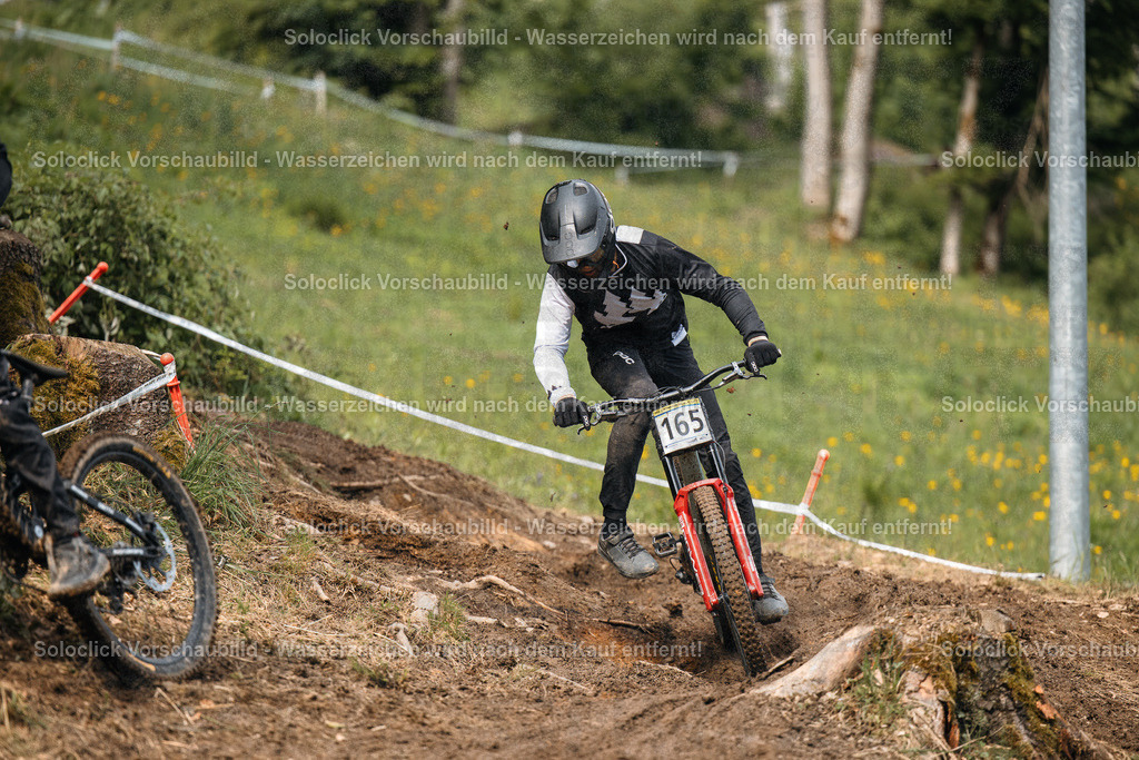 Mountainbiker in Aktion | Ein Mountainbiker rast einen staubigen, kurvigen Trail hinunter. Er trägt einen schwarzen Helm und eine schwarze Jacke mit dem kanadischen Flaggenmuster. Er trägt eine Nummer 165 auf seinem Rücken. Der Trail ist von grünen Bäumen und Büschen gesäumt. Der Himmel ist blau und bewölkt. - Realisiert mit Pictrs.com