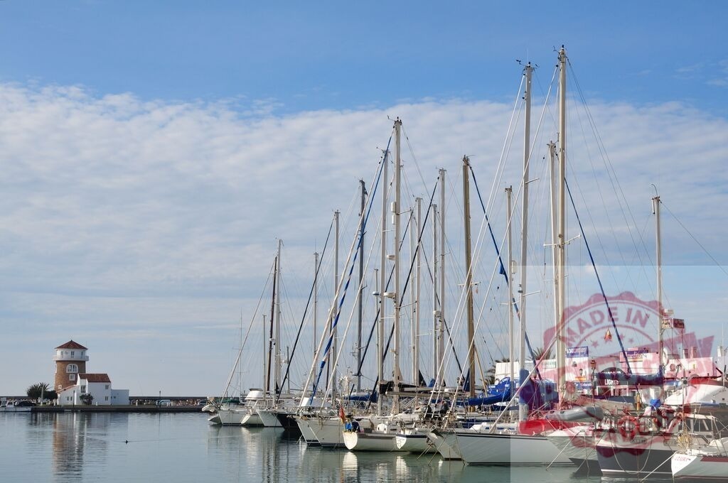 Sailboats moored in marina with lighthouse tower | Boats docked in port, resting near a lighthouse against a cloudy sky - Realisiert mit Pictrs.com