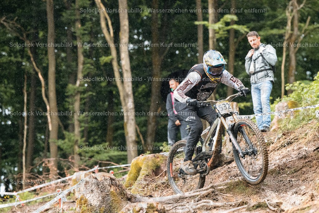 Mountainbike-Abenteuer im Regen | Ein Mountainbiker rast einen steilen, bewaldeten Hang hinunter. Der Fahrer trägt einen schwarzen Helm mit einem weißen Logo, eine dunkle Jacke, schwarze Handschuhe und schwarze Mountainbike-Hosen. Er fährt ein Mountainbike mit schwarzen Reifen und einem weißen Nummernschild mit der Nummer 532. Im Hintergrund sind weitere Bäume und ein Mann in einem grauen Hoodie zu sehen, der den Fahrer beobachtet. Es regnet leicht, was die Szene feucht und dynamisch erscheinen lässt. - Realisiert mit Pictrs.com