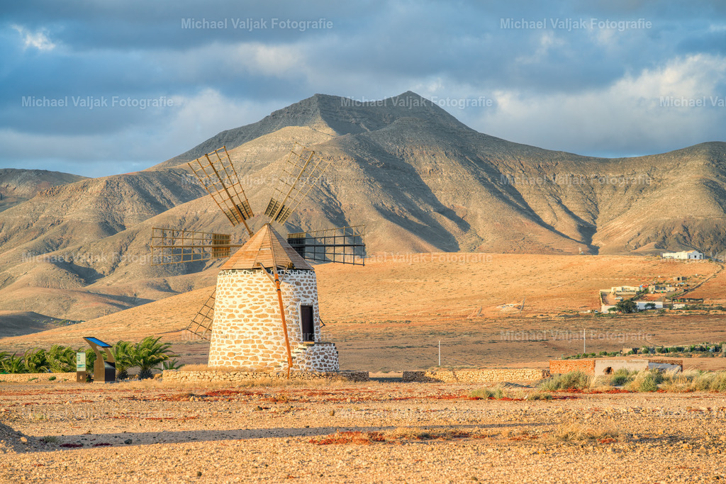 Windmühle in Tefía auf Fuerteventura | Die Windmühle von Tefía steht im warmen Licht des Abends, ihr Holz und Mauerwerk leuchten in sanften Goldtönen. Hinter ihr zeichnen sich die Berge klar gegen den Himmel ab, leicht vom rötlichen Abendlicht gefärbt. Ein stiller Moment, in dem Tradition, Landschaft und das sanfte Abendlicht miteinander verschmelzen. - Realisiert mit Pictrs.com