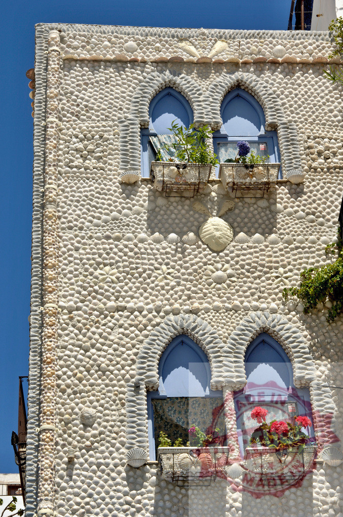 Cadaqués shell house facade with mosaic and blue windows. Provincia de Castellon | Building in Cadaqués decorated with shells, featuring blue windows and flower boxes - Realisiert mit Pictrs.com