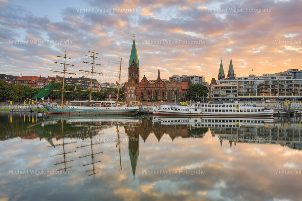 Blick zur Schlachte in Bremen am Morgen | Am Ufer der Weser, kurz nach Sonnenaufgang, zeigt sich ein klarer Blick in Richtung Altstadt. Die ruhige Wasseroberfläche spiegelt die Gebäude entlang des Flusslaufs und verstärkt die Struktur der städtischen Silhouette. Das Segelschiff Alexander von Humboldt liegt im Vordergrund und ergänzt die Szene um ein prägnantes maritimes Detail. Die Aufnahme verbindet städtische Architektur mit der Flusslandschaft in den frühen Morgenstunden. - Realisiert mit Pictrs.com