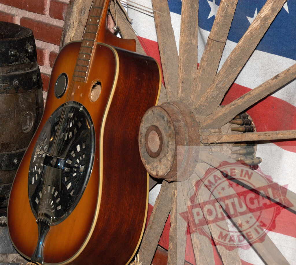Resonator guitar leaning on wagon wheel with american flag and wooden barrel | Resonator guitar leaning on old wagon wheel with american flag and wooden barrel in the background, representing american country music - Realisiert mit Pictrs.com