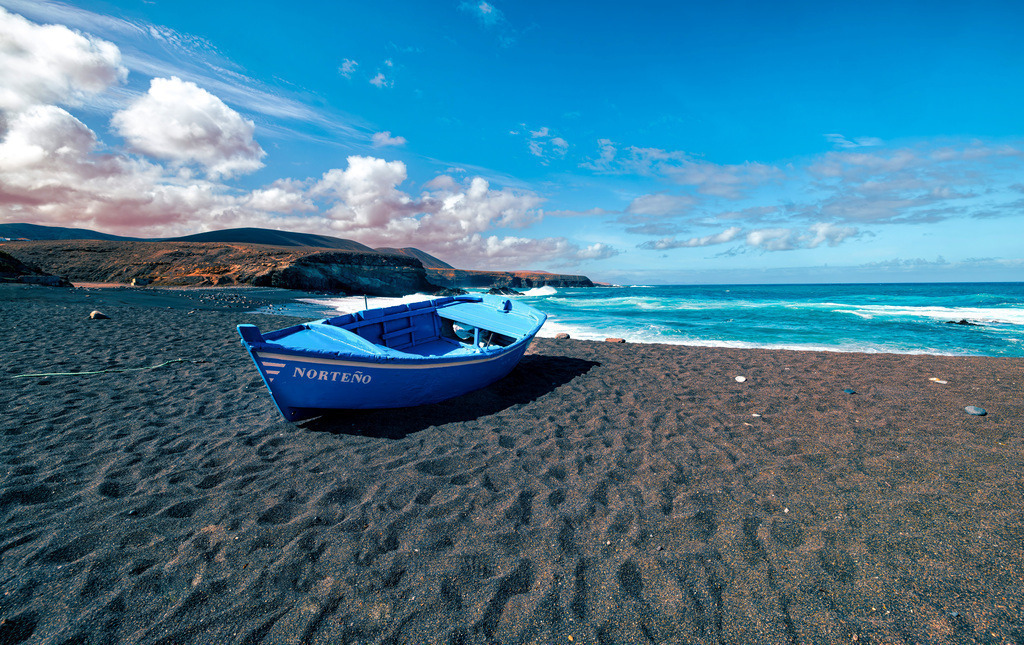 Fischerboot auf schwarzem Vulkansand am Ajuy-Strand auf Fuerteventura | Das Bild zeigt den Ajuy-Strand auf Fuerteventura, Spanien, bekannt für seinen schwarzen Vulkansand. Ort: Ajuy ist ein kleines Fischerdorf an der Westküste von Fuerteventura. Merkmale: Der Strand besteht aus schwarzem Vulkansand und ist ein beliebtes Ausflugsziel mit gemütlichen Restaurants. Sehenswürdigkeiten: In der Nähe befinden sich bekannte Höhlen (Cuevas de Ajuy), die durch den Atlantik geformt wurden. Geschichte: Historisch diente die - Realisiert mit Pictrs.com