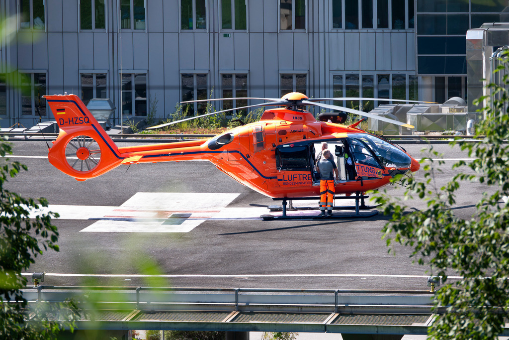 Rettungshubschrauber | Ein leuchtend oranger Rettungshubschrauber der Zivilschutz-Luftrettung (Kennung D-HZSQ) steht startbereit auf dem Landeplatz des Klinikums Garmisch-Partenkirchen. Während das medizinische Personal an der offenen Kabinentür letzte Vorbereitungen trifft, bildet die moderne, silbrig glänzende Fassade des Krankenhauses einen kühlen, technischen Kontrast zum kräftigen Orange der Flugmaschine. - Realisiert mit Pictrs.com