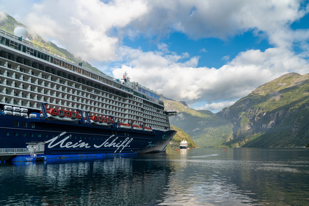 Fjord-Panorama mit Kreuzfahrtschiff | Eine Perspektive vom Wasser auf die Mein Schiff 4 im Geirangerfjord. Die Aufnahme zeigt die beeindruckende Flanke des Kreuzfahrtschiffs am SeaWalk-Anleger, während sich im Hintergrund die Gebirgszüge Norwegens und ein weiteres Schiff in der Ferne abzeichnen.  - Realisiert mit Pictrs.com