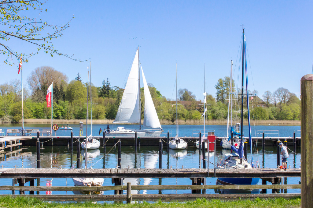 Wandbild: Yachthafen in Kappeln an der Schlei | Dieses Wandbild im Querformat zeigt den Yachthafen in Kappeln an der Schlei im Frühling. Auf der Schlei befindet sich ein Segelboot. Der blaue Himmel ist wolkenlos.  - Realisiert mit Pictrs.com