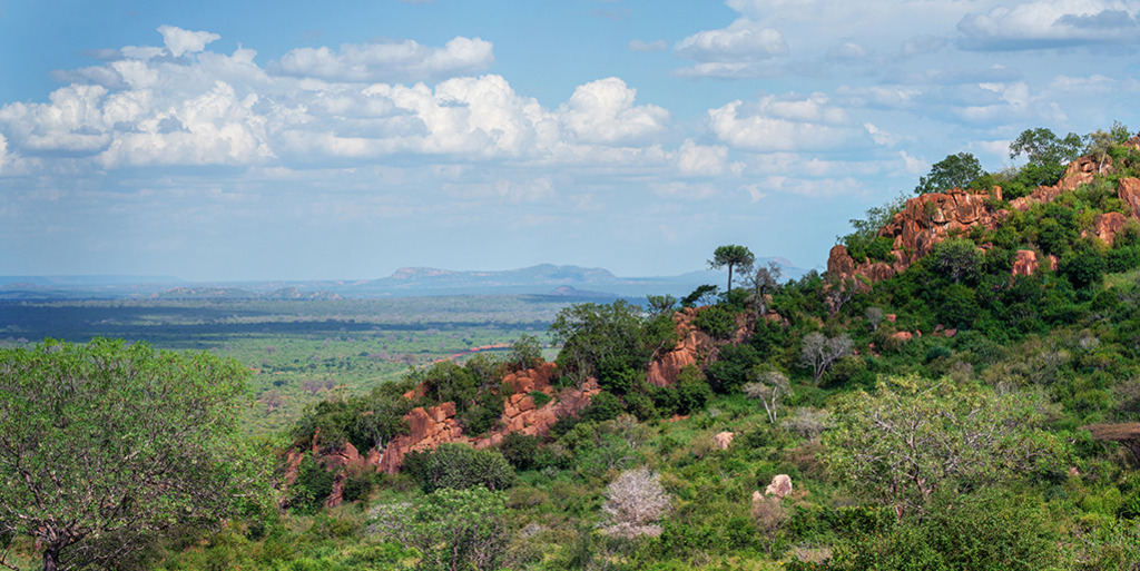 Weite der Savanne (Panorama) | Kenia | Premium-Wandbild im Panorama-Format von justsomewhere.com: Ein spektakulärer Weitblick in Kenia über einen grün bewachsenen, roten Felsrücken hinweg auf die endlose Savanne unter einem weiten Wolkenhimmel. - Realisiert mit Pictrs.com