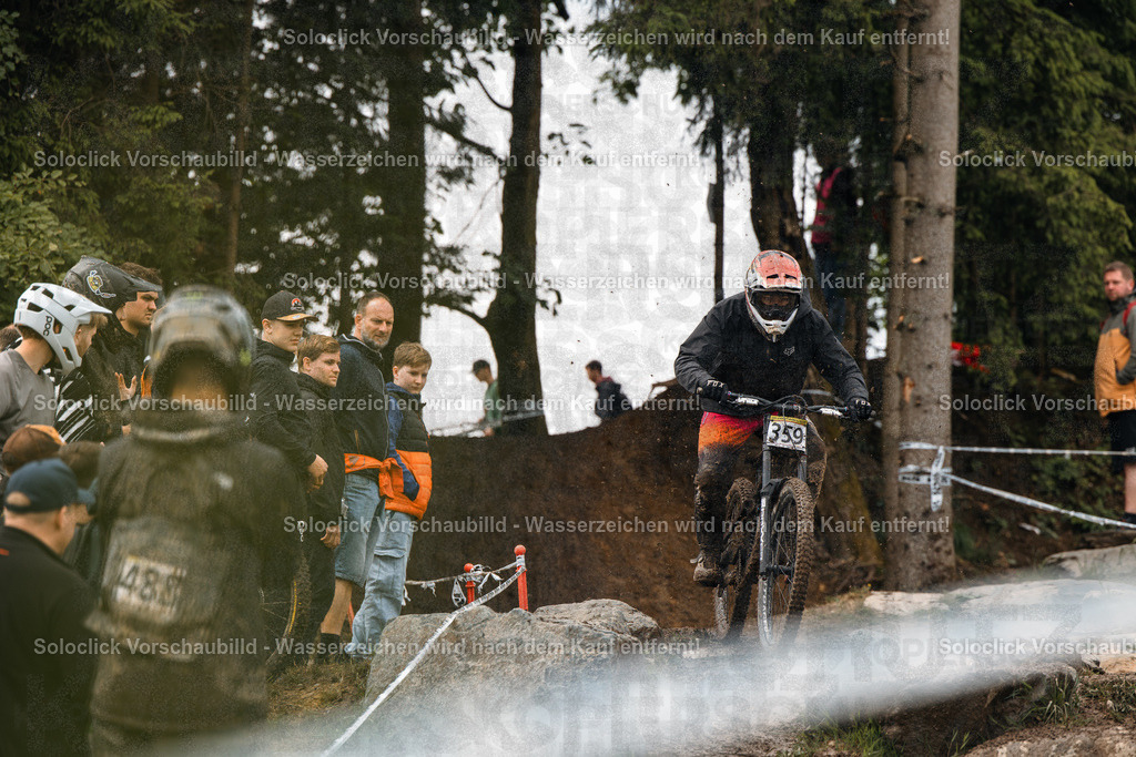 Mountainbike-Action | Ein Mountainbiker in voller Ausrüstung rast auf einem staubigen Trail über einen Sprung. Er trägt einen schwarzen Helm mit orangefarbenen Akzenten, eine dunkle Brille und eine schwarze Jacke. Er trägt auch Handschuhe und Stiefel. Der Trail ist mit Steinen und Wurzeln übersät. Im Hintergrund stehen Zuschauer, die den Fahrer anfeuern. Der Himmel ist bewölkt. - Realisiert mit Pictrs.com