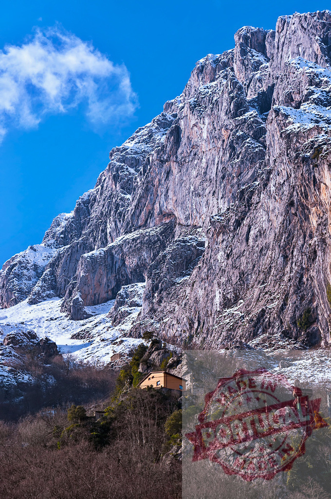 Small mountain house under a huge rocky mountain partially covered with snow | Small mountain house under a huge rocky mountain partially covered with snow, on a sunny day with blue sky and some clouds - Realized with Pictrs.com