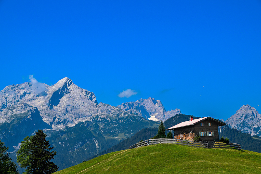 Idylle über den Wolken: Geborgenheit am Haus auf dem Eckbauer | Ein Anblick wie aus einem wunderschönen Traum, der tiefe Sehnsucht und heimatliche Geborgenheit weckt: Auf einer saftig grünen, sonnenbeschienenen Almkuppe thront das urige Haus auf dem Eckbauer, geschützt von einem hölzernen Zaun. Dahinter ragt in atemberaubender Pracht das gewaltige Massiv der Alpspitze und der angrenzenden Wettersteingipfel empor. Die schroffen Felswände zeichnen sich gestochen scharf vor einem makellosen, tiefblauen Sommerhimmel ab. Diese Aufnahme fängt die vollkommene Ruhe und die friedliche Einsamkeit der Alpen perfekt ein – ein Zufluchtsort für die Seele, der die Zeit für einen Moment stillstehen lässt. - Realisiert mit Pictrs.com