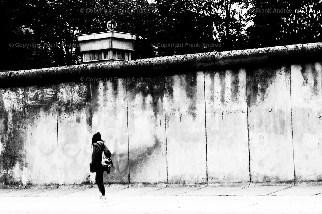 Berliner Mauer | Eine junge Frau geht entlang der Berliner Mauer mit Wachturm. Straßenfotografie aus Berlin in schwarz und weiß. - Realisiert mit Pictrs.com