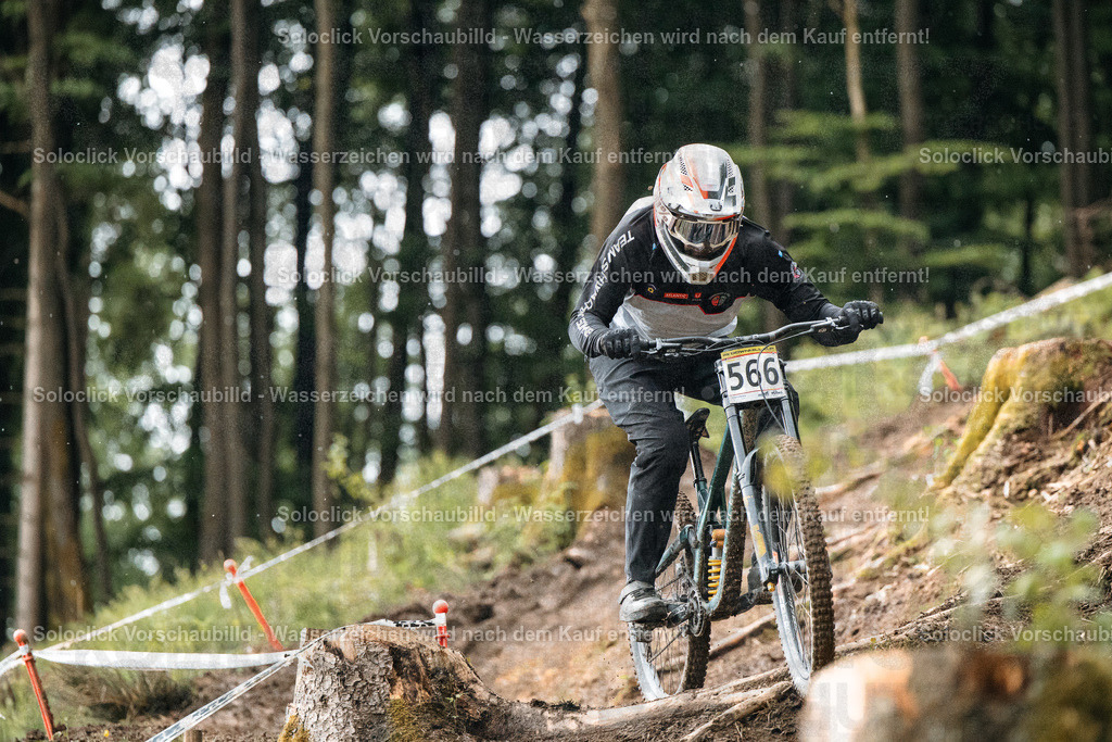 Mountainbiker im Wald | Ein Mountainbiker in voller Ausrüstung rast mit hoher Geschwindigkeit einen staubigen, bewaldeten Kurs hinunter. Er trägt einen schwarzen Helm mit einer roten und weißen Grafik, eine dunkle Brille, eine schwarze Jacke und Hose. Er trägt eine Nummer 566 auf seinem Rücken. Der Kurs ist mit roten und weißen Markierungen versehen und führt durch einen dichten Wald. Der Boden ist staubig und uneben. - Realisiert mit Pictrs.com