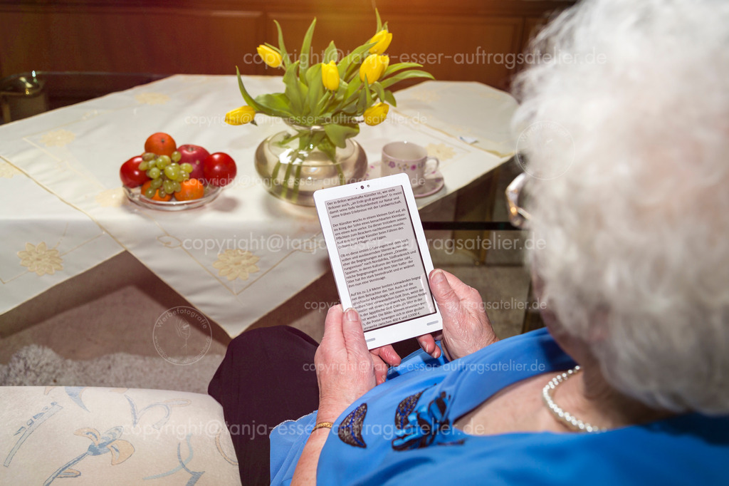 Seniorin benutzt E-Book D | Eine 80 jährige Seniorin sitzt auf ihrer Couch und liest auf einem E Reader. Deutscher Text ist in komfortabler Größe lesbar. Text ist freigegeben. Ansicht über die Schulter. 