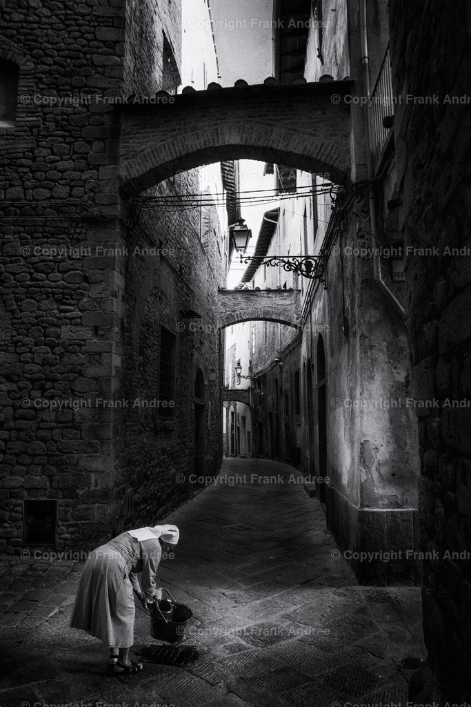 Nonne in Pistoia | Eine Nonne leert einen Eimer in einer engen Gasse in der Altstadt von Pistioa Toskana. Straßenfotografie aus Italien in schwarz / weiß. - Realisiert mit Pictrs.com
