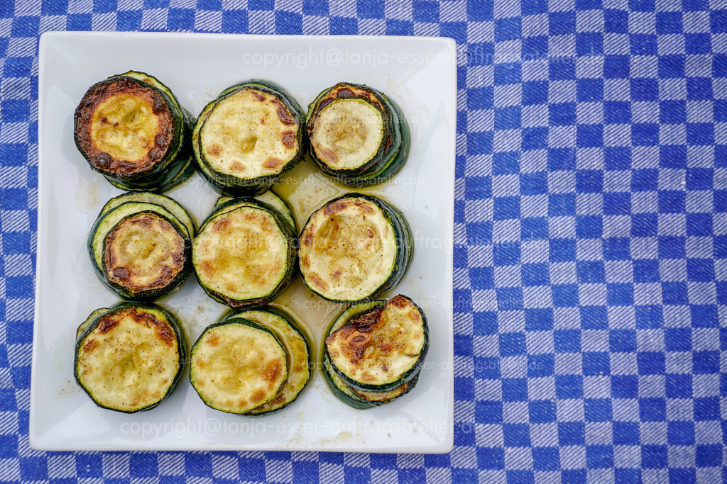 Fried zucchini slices D | Ein weißer Teller mit gebratenen Zucchini Scheiben im Bratensaft. Das Gemüse steht auf einem blauen Küchenhandtuch und wurde von oben fotografiert mit Copy Space. 