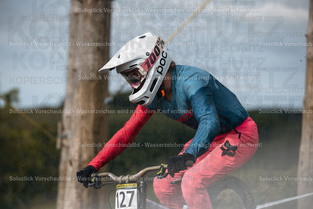 Mountainbiker im Rennen | Ein Mountainbiker in voller Montainbike-Ausrüstung rast einen staubigen, kurvigen Pfad hinunter. Er trägt einen weißen Helm mit der Aufschrift 'POC' und ein blaues Trikot mit einem roten Tuch. Seine Hose ist rot. Er hält ein Mountainbike mit roten und schwarzen Komponenten. Der Hintergrund besteht aus Bäumen und Erde. - Realisiert mit Pictrs.com