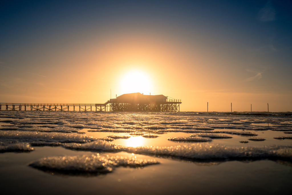 Sonnenuntergang in St. Peter Ording | Auf diesem Bild ist eine malerische Szene am Strand von St. Peter-Ording zu sehen, in der die berühmten Pfahlbauten bei tiefstehender Sonne im Hintergrund erscheinen. Das goldene Licht der untergehenden Sonne taucht die Szenerie in warme Töne und spiegelt sich sanft im feuchten Sand und den kleinen Wasserpfützen des Wattenmeers. Die Struktur der Holzpfähle und die Stille der Umgebung verleihen dem Bild eine friedliche und ruhige Atmosphäre, die an die natürliche Schönheit der Nordseeküste erinnert.