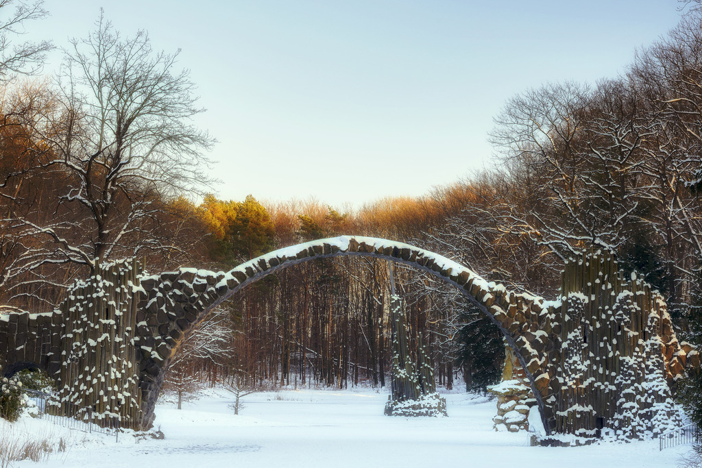 Teufelsbrücke, Deutschland | Das Bild zeigt die Rakotzbrücke, oft auch Teufelsbrücke genannt,  im winterlichen Azaleen- und Rhododendronpark Kromlau. Der Schnee und die letzten Sonnenstrahlen des Tages, die die Baumwipfel zum Strahlen bringen, tauchen das märchenhafte Bauwerk in ein mystisches Licht.Unsere Empfehlung: Alu-Dibond oder Acrylglas - Realisiert mit Pictrs.com