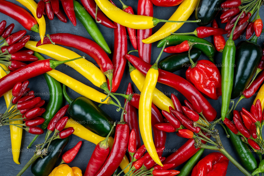 Leuchtende Chili-Vielfalt in Rot, Gelb und Grün | Zahlreiche Chilischoten in verschiedenen Formen und Farben bilden ein buntes Mosaik auf neutralem Hintergrund. Rote Chilisträuße treffen auf einzelne gelbe, schwarze und grüne Schoten unterschiedlicher Größen. Die natürliche Anordnung zeigt die Diversität dieser Gewürzpflanzen eindrucksvoll. Lange schmale Schoten wechseln sich mit rundlichen Exemplaren ab, während die intensiven Farben von feurigem Rot über sonniges Gelb bis zu geheimnisvollem Schwarz reichen. Diese Food-Fotografie demonstriert die kulinarische Bandbreite scharfer Küchengewürze perfekt. - Realisiert mit Pictrs.com