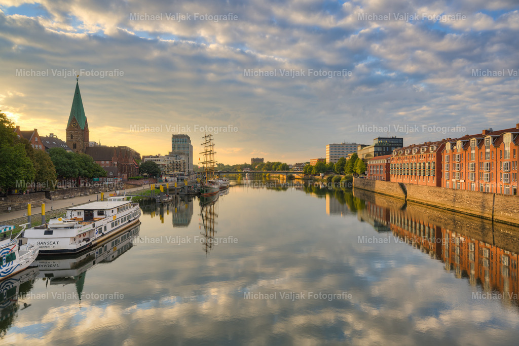 Blick über die Weser in Bremen | Ein ruhiger Morgen an der Weser in Bremen: Die ersten Sonnenstrahlen tauchen die Häuser auf der rechten Flussseite in ein warmes Licht und lassen die Fassaden in sanften Farbtönen erscheinen. Links im Bild erstreckt sich die Altstadt mit der historischen Schlachte, wo Gastronomie und Spazierwege das Ufer säumen. Das Segelschiff Alexander von Humboldt liegt gut sichtbar am Kai und ergänzt die Szene um ein markantes maritimes Detail. Die Komposition verbindet städtisches Leben mit der Flusslandschaft in den frühen Stunden des Tages. - Realisiert mit Pictrs.com