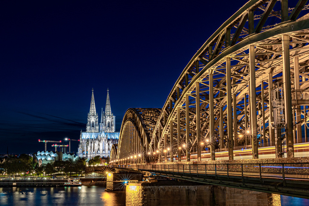 City of Lights – Cologne Connection | Under the deep blue evening sky, Cologne’s Cathedral rises in luminous grace while the Hohenzollern Bridge glows with the rhythm of passing trains. A meeting of history and motion — where Gothic spires and steel arcs echo the pulse of an ever-moving city. - Realisiert mit Pictrs.com