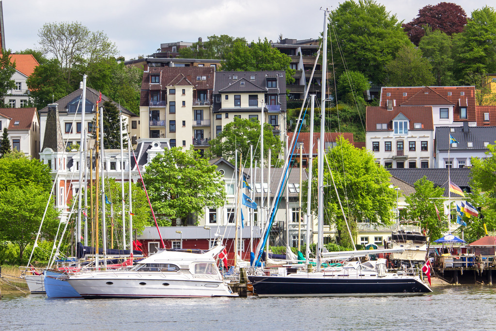 Wandbild: Hafen in Flensburg im Frühling | Dieses Wandbild im Querformat zeigt den Hafen in Flensburg im Frühling. Zwischen den Gebäuden auf der anderen Uferseite ist schönes Frühlingsgrün zu sehen. Am blauen Himmel sind zahlreiche Wolken zu sehen.  - Realisiert mit Pictrs.com