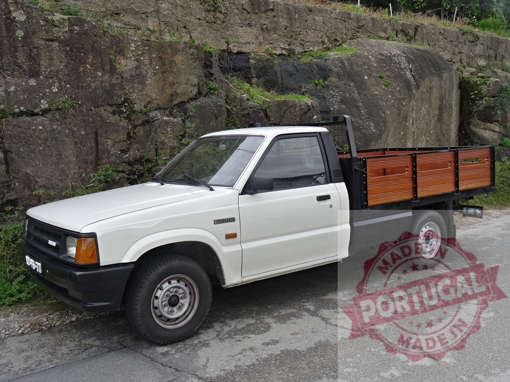 White Mazda B2500 flatbed truck with wooden cargo bed | White Mazda B2500 pickup truck featuring a wooden flatbed cargo body parked outdoors - Realized with Pictrs.com