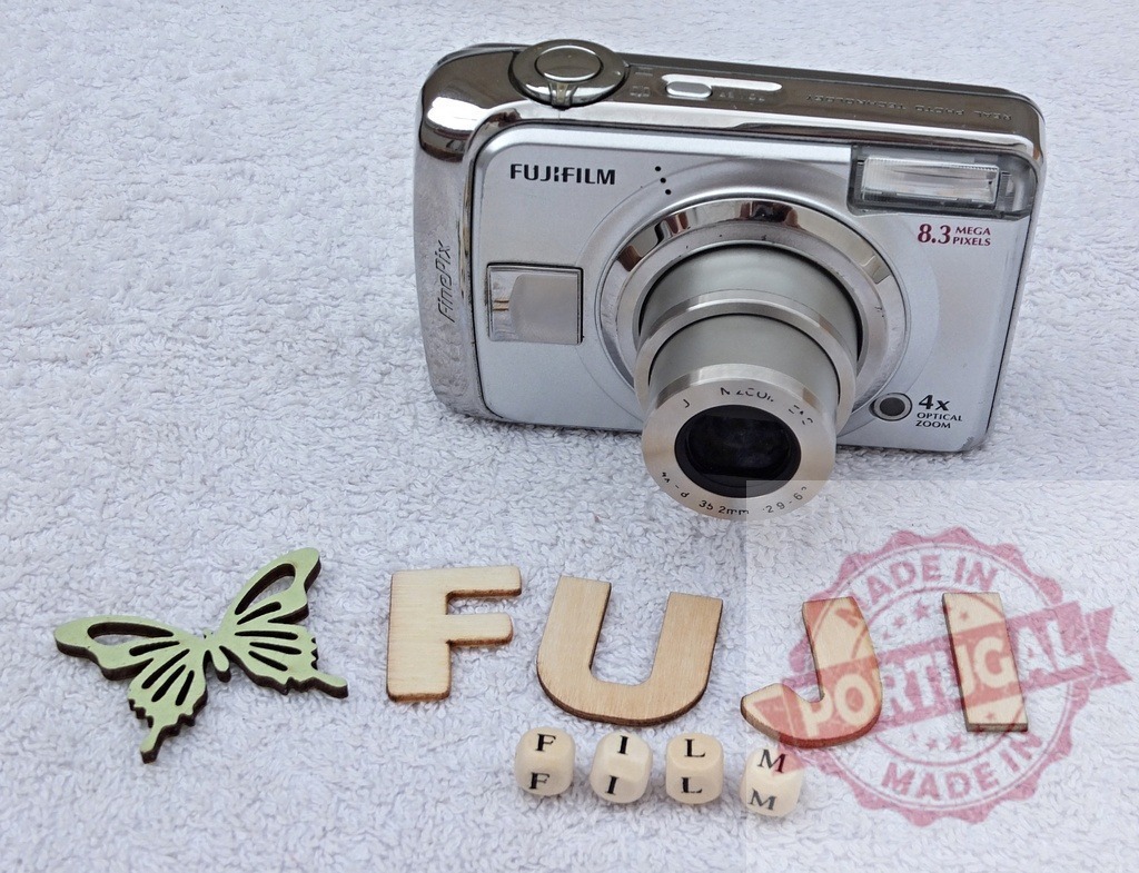 Fujifilm FinePix digital camera with wooden letters | Fujifilm FinePix digital camera, wooden letters spelling out FujiFilm, and a butterfly decoration on a white background - Realized with Pictrs.com