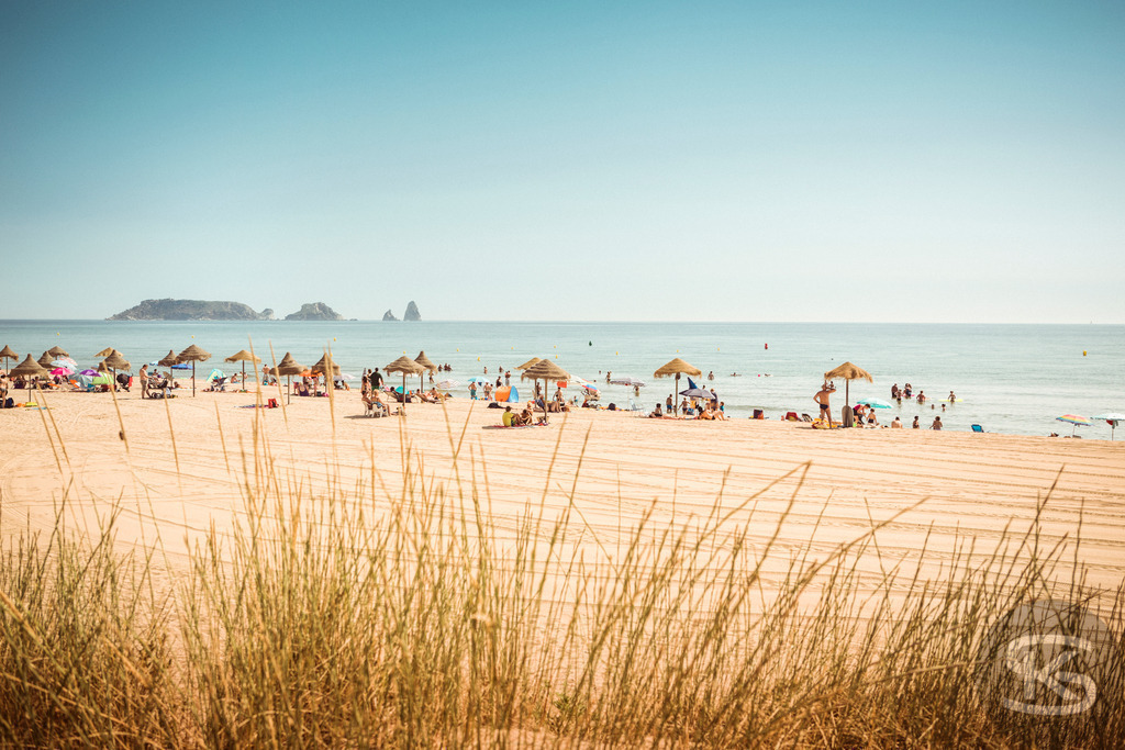 Sommerlicher Sandstrand mit Urlaubern und Meerblick | Ein lebhaftes und sonniges Bild eines Sandstrandes voller Urlauber. Strohschirme säumen den Strand, Menschen entspannen und schwimmen im kühlen Meerwasser. Im Hintergrund sind kleine Inseln sichtbar. Die Szene fängt die fröhliche und entspannte Sommer-Atmosphäre eines perfekten Strandurlaubs ein. - Realisiert mit Pictrs.com