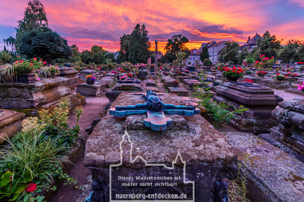 Nürnberger Stadtbild | Der Johannisfriedhof in Nürnberg bei Sonnenuntergang. Im Vordergrund ist ein altes Grab mit einem verwitterten Grabkreuz, das den gekreuzigten Jesus zeigt. Der Himmel ist in Orange- und Violetttönen gefärbt. Die Gräber sind mit Blumen geschmückt, - Realisiert mit Pictrs.com
