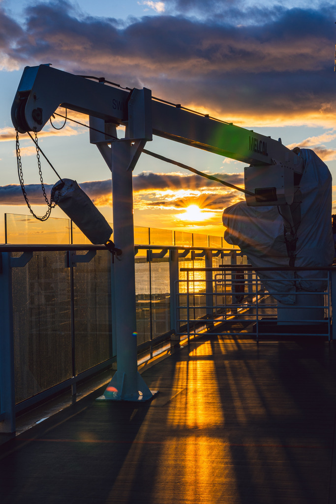 Sonnenuntergang mit Kran | Abendstimmung auf hoher See: Ein Kran an Deck steht im Kontrast zu einem wolkenverhangenen Himmel, während die Sonne am Horizont für einen atemberaubenden Farbakzent sorgt. - Realisiert mit Pictrs.com