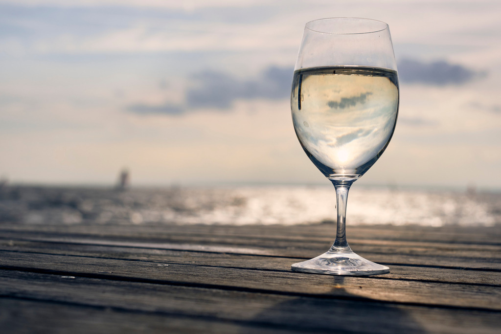 Ein Glas Weisswein auf einem Holzsteg am Neusiedlersee | Burgenland, Austria - October 05, 2021: Ein Glas Weisswein auf einem Holzsteg stehend am Neusiedlersee, im Hintergrund das Wasser und ein Segelboot. - Realisiert mit Pictrs.com