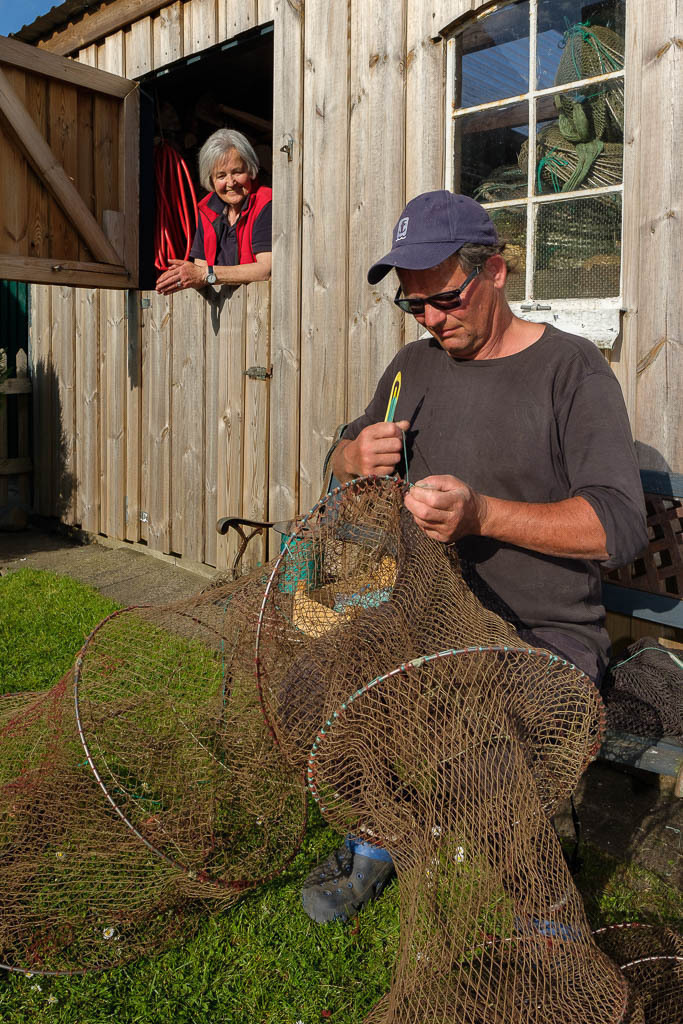 zeitenwende-farbe-01-08 | Matthias Nanz bei der Reparatur einer Reuse auf seinem Hofplatz unter den wachsamen Augen seiner Mutter Lisa Nanz. Die Reparatur der Netze und Reusen ist eine ständige Herausforderung für die Fischer vom Holm. Mit einer Netznadel werden kleine Löcher gestopft, durch die Fische sonst leicht entkommen könnten. - Realisiert mit Pictrs.com