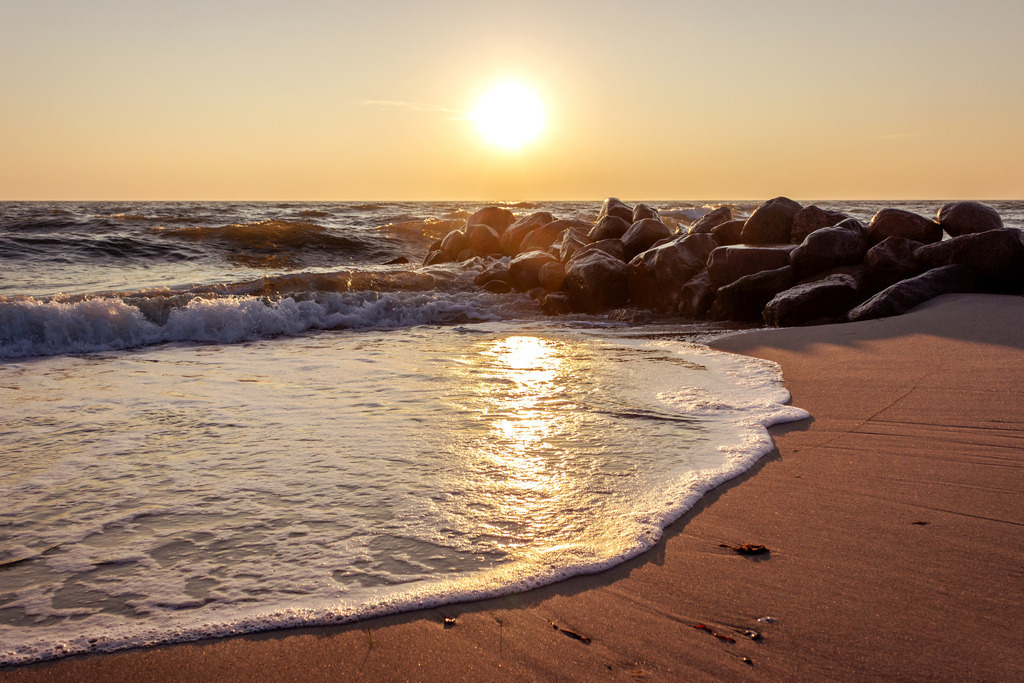 Wandbild: Sonnenaufgang an der Ostsee | Dieses Wandbild im Querformat zeigt einen schönen Sonnenaufgang an der Ostsee. Die morgendliche Sonne erzeugt zahlreiche Reflexionen auf einer auslaufenden Welle am Sandstrand. Von der rechen Seite ragt eine Steinmole ins Wandbild - Realisiert mit Pictrs.com
