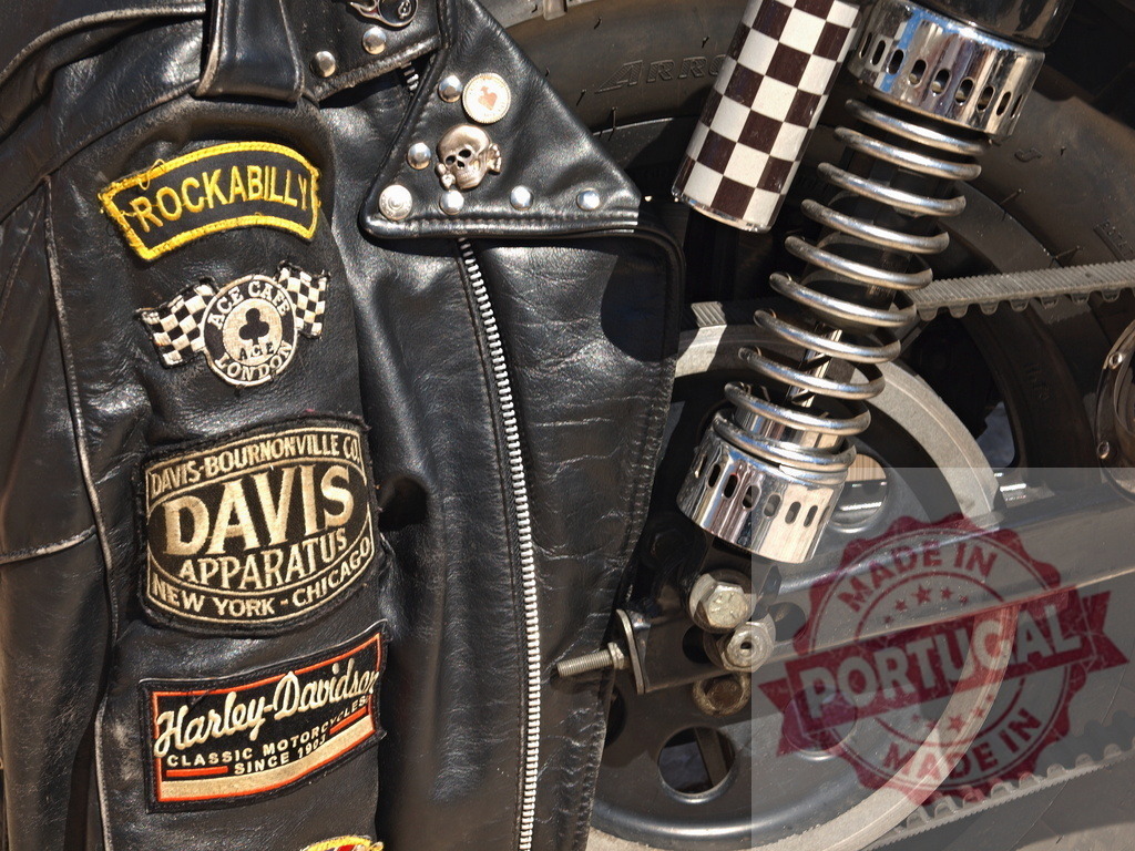 Leather jacket with Rockabilly and Harley-Davidson patches on motorcycle | Black leather jacket displaying various patches and pins alongside a motorcycle suspension - Realisiert mit Pictrs.com