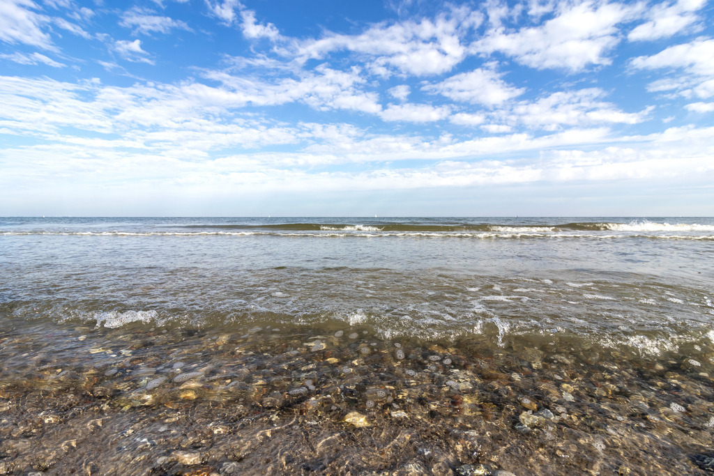 Wandbild: Wellen am Ostseestrand in Falshöft | Dieses Wandbild im Querformat zeigt Wellen am Strand in Falshöft. Durch das klare Wasser sind Steine auf dem Meeresboden zu sehen. Am blauen Himmel sind einige sommerliche Wolken zu sehen.  - Realisiert mit Pictrs.com