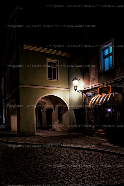 Stille Gasse bei Nacht | Atmosphärische Nachtaufnahme einer alten Gasse mit Kopfsteinpflaster, beleuchteter Laterne und blauem Fensterlicht. Die starken Kontraste, das warme Kunstlicht und die dunkle Umgebung erzeugen eine ruhige, geheimnisvolle und leicht filmische Stimmung. - Realisiert mit Pictrs.com