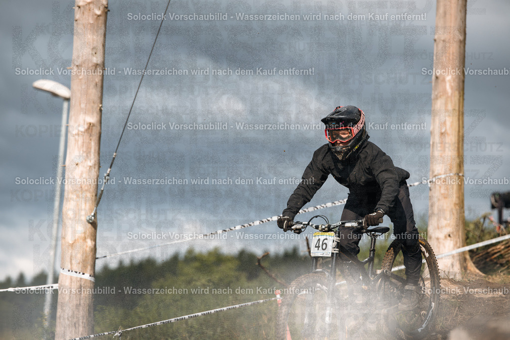 Mountainbike-Action im Gelände | Das Bild zeigt einen Mountainbiker in voller Ausrüstung, der einen staubigen Pfad mit hoher Geschwindigkeit durchquert. Der Fahrer trägt einen schwarzen Helm mit einer gelben und roten Markierung sowie eine dunkle Schutzkleidung. Der Hintergrund besteht aus Bäumen und einem Himmel mit dunklen Wolken. Staub wirbelt um das Fahrrad auf, was die Geschwindigkeit des Fahrers verdeutlicht. - Realisiert mit Pictrs.com