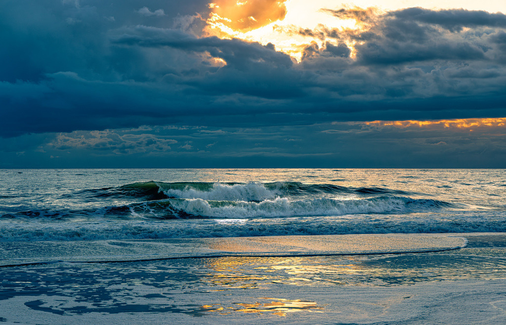 Stimmungsvolle Meerlandschaft am Strand von Westerland,Sylt | Das Bild zeigt eine Meereslandschaft bei Sonnenuntergang am Strand von Westerland auf SyltDetails der SzeneHimmel: Dunkle, schwere Wolken verdecken den Großteil des Himmels, während ein goldener Lichtschimmer im oberen Zentrum durchbricht.Meer: Eine breite Welle bricht sich im mittleren Bildbereich und erzeugt weißen Gischt.Vordergrund: Der nasse Sandstrand reflektiert das warme, goldene Licht des Himmels. - Realisiert mit Pictrs.com