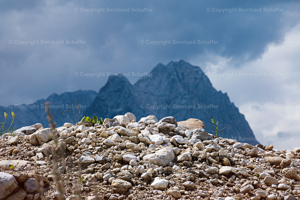 Steinhuegel vor Bergmassiv | Steiniger Hügel mit Pflanzen vor einer Bergkette und Gewitterwolken am Himmel. - Realisiert mit Pictrs.com