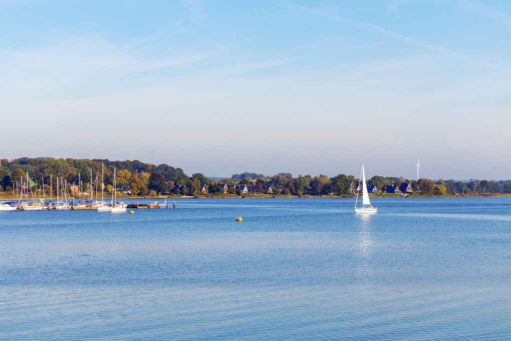Wandbild: Yachthafen und Segelboot in Lindaunis | Dieses Wandbild im Querformat zeigt ein Segelboot auf der Schlei in Lindaunis. Auf der linken Seite ist der Yachthafen in Lindaunis zu sehen. Am blauen Himmel befinden sich einige Schleierwolken. Das Segelboot spiegelt sich schön auf der Wasseroberfläche der Schlei.  - Realisiert mit Pictrs.com