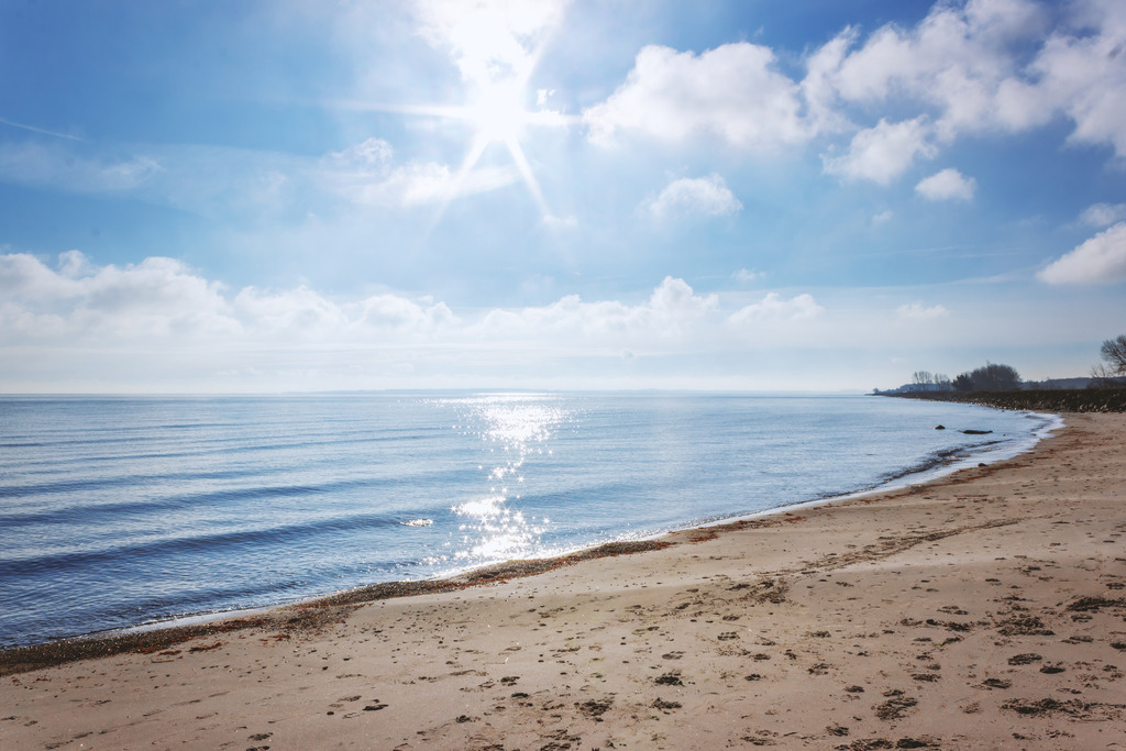 Wandbild: Funkelndes Meereslicht | Ein Moment von purer Klarheit und nordischer Frische. Dieses Wandbild fängt das erste, helle Licht eines neuen Tages an der Küste ein. Der Blick schweift über den feinen Sandstrand und das ruhig pulsierende Blau des Meeres bis hin zur sanften Silhouette der fernen Uferlinie. Die kristallklare Luft und das glitzernde Sonnenlicht auf den Wellen verleihen diesem Motiv eine belebende Tiefe. Ein minimalistisches Kunstwerk, das die weite Ruhe und die frische Energie eines Morgens am Meer direkt in Ihr Zuhause bringt. - Realized with Pictrs.com