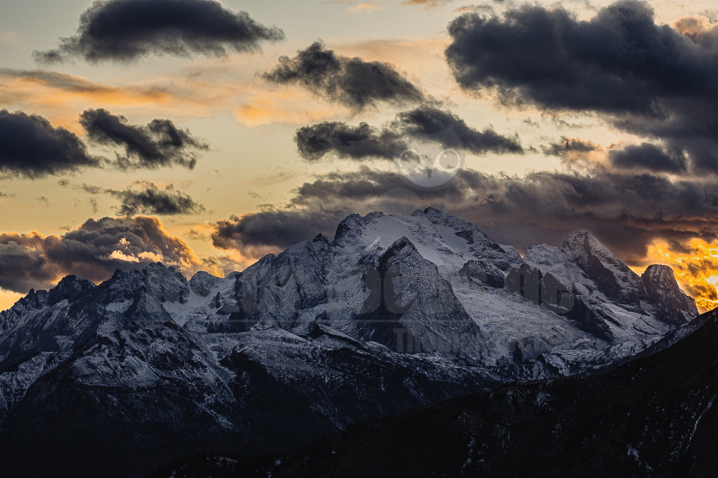 R6NF7217_20251005 | Ein atemberaubender Querformat-Blick auf eine majestätische Bergkette, deren Gipfel mit frischem Schnee bedeckt sind. Der Himmel ist von dunklen, dramatischen Wolken durchzogen, die von den warmen, goldenen und orangefarbenen Tönen des Sonnenuntergangs durchbrochen werden. Die tief stehende Sonne taucht die Szene in ein dramatisches Licht, das die schroffen Konturen der Berge hervorhebt und einen starken Kontrast zu den dunklen Schatten im Vordergrund bildet. Die Szene strahlt eine ruhige, aber kraftvolle Atmosphäre aus, die typisch für die hochalpine Landschaft der Dolomiten ist, insbesondere die Marmolada. - Realisiert mit Pictrs.com